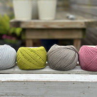 Four balls of wool in silvery grey, rich earthy lime green, medium grey, and medium soft pink. 