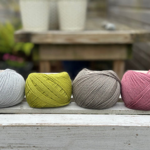 Four balls of wool in silvery grey, rich earthy lime green, medium grey, and medium soft pink. 