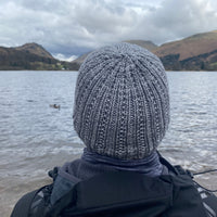 A person looking out over a lake towards mountains. The person is wearing a grey knitted hat with textured stitches running up the height of the hat and gathering at the crown