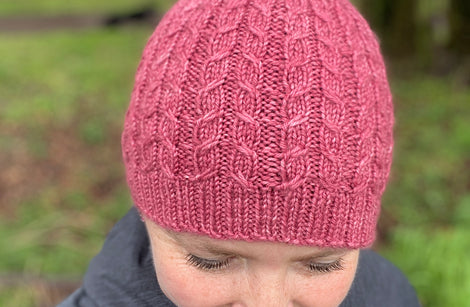 Victoria looking towards the floor wearing a pink knitted hat. The hat has a cable pattern and a small bobble on the crown