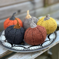 Four knitted pumpkins on a plate - the colours of them are rusty brown, charcoal, yellow, light brown, and orange.