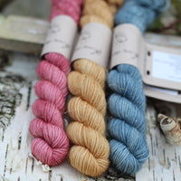 Three mini skeins of yarn with gold sparkle running through it. From left to right: a purpley red skein, a golden brown skein and a blue skein