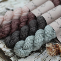 Four mini skeins of yarn. From left to right: a beige skein, a brown skein, a dark grey skein and a grey skein