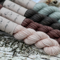 Four mini skeins of yarn. From left to right: a beige skein, a brown skein, a dark grey skein and a grey skein