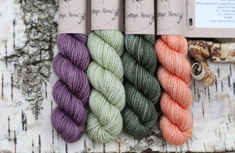 Four mini skeins of yarn. From left to right: a purple skein, a pale green skein, a dark green skein and an orange skein 