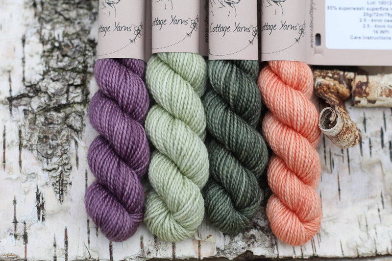 Four mini skeins of yarn. From left to right: a purple skein, a pale green skein, a dark green skein and an orange skein 