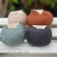 Four balls of yarn in two rows of two balls. On the left there is a beige ball and a blue-green ball. On the right there is a reddish brown ball and a dark grey ball