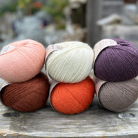 Six balls of yarn arranged in two rows of three balls. The top row is peachy orange, cream and dark purple. The bottom row is reddish brown, orange and grey