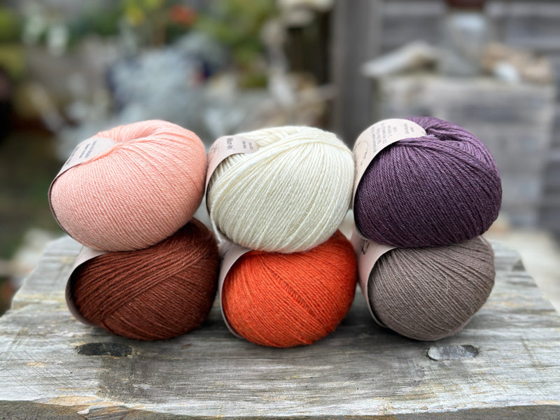 Six balls of yarn arranged in two rows of three balls. The top row is peachy orange, cream and dark purple. The bottom row is reddish brown, orange and grey