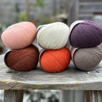 Six balls of yarn arranged in two rows of three balls. The top row is peachy orange, cream and dark purple. The bottom row is reddish brown, orange and grey