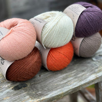 Six balls of yarn arranged in two rows of three balls. The top row is peachy orange, cream and dark purple. The bottom row is reddish brown, orange and grey