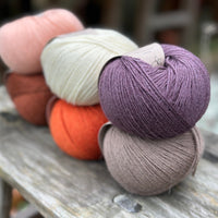 Six balls of yarn arranged in two rows of three balls. The top row is peachy orange, cream and dark purple. The bottom row is reddish brown, orange and grey