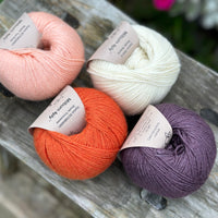 Four balls of yarn. From left to right there is a peachy pink ball, an orange ball, a cream ball and a dark purple ball.