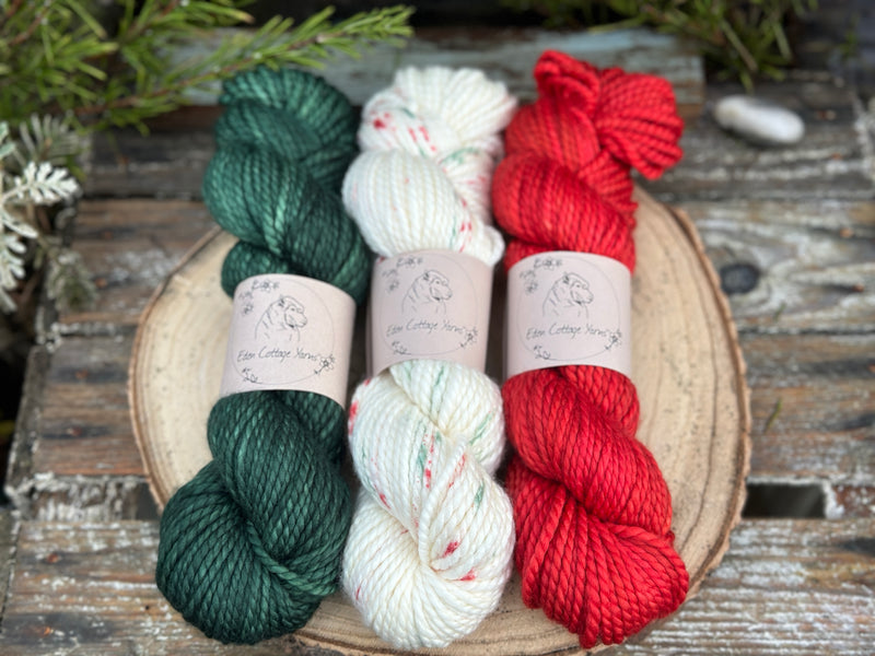 Three skeins of yarn. From left to right: a dark green skein, a cream skein with green and red speckles and a bright red skein
