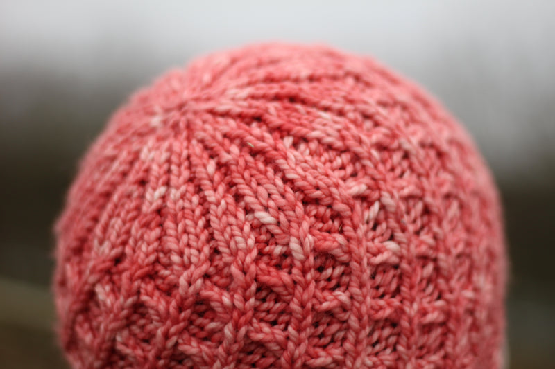 The crown of a bright pink beanie hat. The all over diamond cable stitch pattern gathers at  the crown
