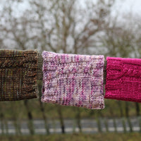 Three cowls draped over a wooden fence. From left to right the cowls are variegated brown, variegated pink and purple, and rich pink. Each cowl has a single cable visible 