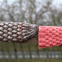 Two hats resting on a wooden fence. On the left hand side of the image is a two tone cable lattice in cream and beige with a dark grey and red contrast. On the right hand side is a bright pink hat in the same cable lattice design but one colour