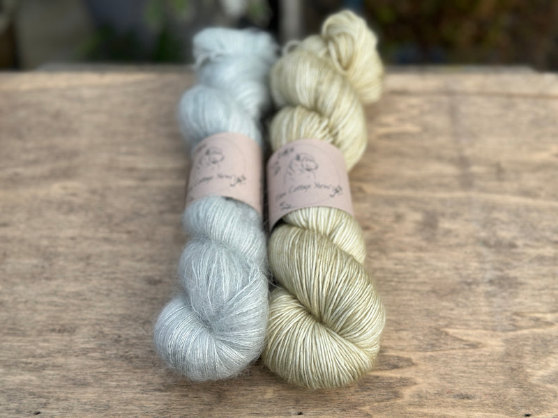 Two skeins of yarn - one is a pale blue fluffy laceweight skein and one is a pale green skein.