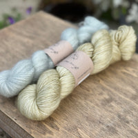 Two skeins of yarn - one is a pale blue fluffy laceweight skein and one is a pale green skein.