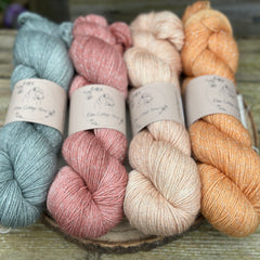 Four skeins of yarn with silver sparkle running through it. From left to right: a grey skein, a pink skein, a pale orange skein and an orange skein