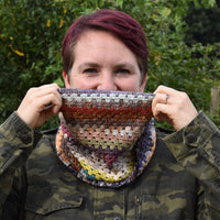 Victoria looking into the camera and pulling a multicoloured cowl up from her neck over her mouth. The cowl features granny stripes in many different colours 