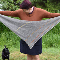 Victoria holding a triangular grey shawl outstretched while a small black dog looks on. The shawl is grey with thin stripes of contrasting colours