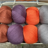 Eight balls of yarn in four pairs. From left to right the colourways are red-brown, purple, orange and grey