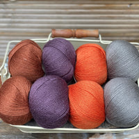 Eight balls of yarn in four pairs. From left to right the colourways are red-brown, purple, orange and grey