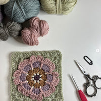 A flat lay including part used balls of yarn in the top left corner, a granny square is shown at the bottom of the image using the same colours of yarn. In the bottom right corner is a crochet hook, a small pair of scissors and a stitch marker.
