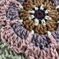 Close up of a granny square. Colours used in the design are dark purple, cream, brown, pale purple, pink and pale green.