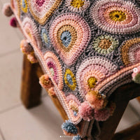 A crochet wrap made in circular motifs of many colours is draped over a wooden stool. The shawl has many multicoloured pom poms along the edges.