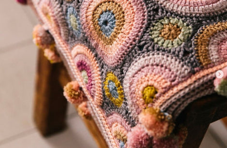 A crochet wrap made in circular motifs of many colours is draped over a wooden stool. The shawl has many multicoloured pom poms along the edges.