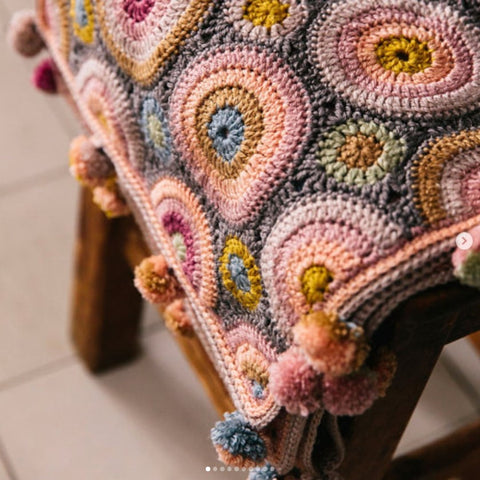 A crochet wrap made in circular motifs of many colours is draped over a wooden stool. The shawl has many multicoloured pom poms along the edges.