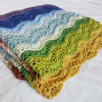 A folded crochet blanket featuring a striped ripple stitch pattern. The blanket uses all twenty colours of Milburn DK but only some of these colours are visible. The pattern is Elysium by Sarah Alderson
