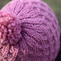 Close up image of a pink hat with cable lattice pattern and a pink pom pom