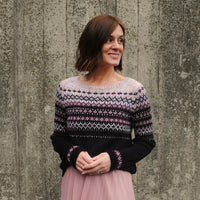 Rachel stood in front of a wall wearing a colourwork jumper with intricate detailing to the yoke, sleeves and body. The colours in the jumper are beige, light pink, purple and black.