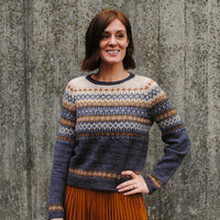 Rachel stood in front of a wall wearing a colourwork jumper with intricate detailing to the yoke, sleeves and body