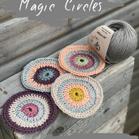 Four crocheted circles are sat on a wooden shelf, in colours of aubergine, beige, peach, pink, silver, yellow, and sage green. They are next to a steely grey ball of Milburn DK yarn. 