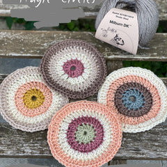 Four crocheted circles are sat on a wooden bench, in colours of brown, beige, peach, pink, cream, yellow, and sage green. They are next to a steely grey ball of Milburn DK yarn. 