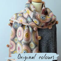 A mannequin wearing a plain grey jumper is displaying a crocheted scarf or wrap. It has circle motifs, as well as smaller semi-circles, and cute little pompoms around the edge. The palette is a grey background with the circles in a variety of dusky pastel colours. Text at the top says 'Magic Circles' and text at the bottom says 'Original colours'.