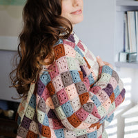 A person with long brown hair wearing a patchwork granny square jacket