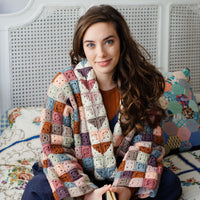 A person with long brown hair sat on a bed wearing a patchwork granny square jacket