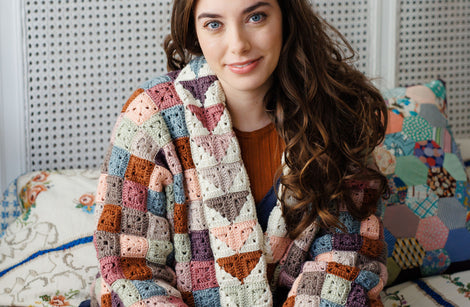 A person with long brown hair sat on a bed wearing a patchwork granny square jacket