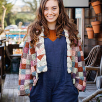 A person with long brown hair wearing a patchwork granny square jacket over blue dungarees