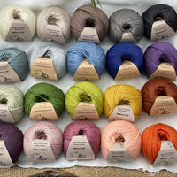 Twenty balls of yarn are arranged in four rows of five balls. The yarn is resting on a white piece of fabric in a jute lined wooden box. From left to right, the colours in the top row are Natural, Wicker, Compost, Steel and Charcoal. The colours in the second row are Catmint, Rain, Estuary, Night Sky and Damson. The colours in the third row are Thyme, Moss, Fern, Harvest Gold and Rust. The colours in the bottom row are Black Tulip, Althaea, Bramble, Tea Rose and Crocosmia.