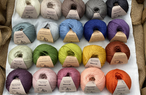 Twenty balls of yarn are arranged in four rows of five balls. The yarn is resting on a white piece of fabric in a jute lined wooden box. From left to right, the colours in the top row are Natural, Wicker, Compost, Steel and Charcoal. The colours in the second row are Catmint, Rain, Estuary, Night Sky and Damson. The colours in the third row are Thyme, Moss, Fern, Harvest Gold and Rust. The colours in the bottom row are Black Tulip, Althaea, Bramble, Tea Rose and Crocosmia.