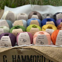 19 balls of yarn are arranged in three rows of five balls and one row of four in the foreground. The yarn is resting on a white piece of fabric in a jute lined wooden box. From left to right, the colours in the top row are Natural, Wicker, Compost, Steel and Charcoal. The colours in the second row are Catmint, Rain, Estuary, Night Sky and Damson. The colours in the third row are Thyme, Moss, Fern, Harvest Gold and Rust. The colours in the bottom row are Black Tulip, Bramble, Tea Rose and Crocosmia.