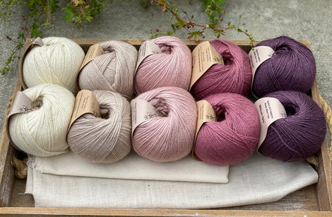 10 balls of yarn are sat in a wooden tray, there are two balls of each colour. The colours from left to right are Natural, Wicker, Althaea, Bramble and Black Tulip. The yarns create a fade effect from natural cream, through pink to dusky purple.