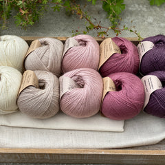 10 balls of yarn are sat in a wooden tray, there are two balls of each colour. The colours from left to right are Natural, Wicker, Althaea, Bramble and Black Tulip. The yarns create a fade effect from natural cream, through pink to dusky purple.