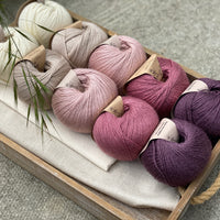 10 balls of yarn are sat in a wooden tray, there are two balls of each colour. The colours from left to right are Natural, Wicker, Althaea, Bramble and Black Tulip. The yarns create a fade effect from natural cream, through pink to dusky purple.